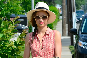 Woman in hat and sunglasses, wearing a patterned sleeveless shirt, outdoors near a car and greenery.