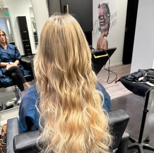 Woman with long, wavy blonde hair sitting in a salon chair, back to the camera.