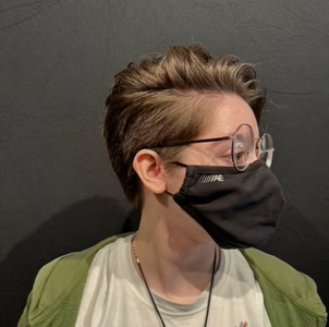 Person with short, wavy hair wearing glasses and a black face mask, against a dark background, in a green jacket.