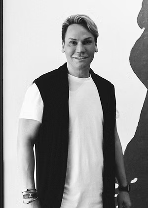 Man smiles, wearing a black vest over a white shirt. Stands against a white wall with a dark shape.