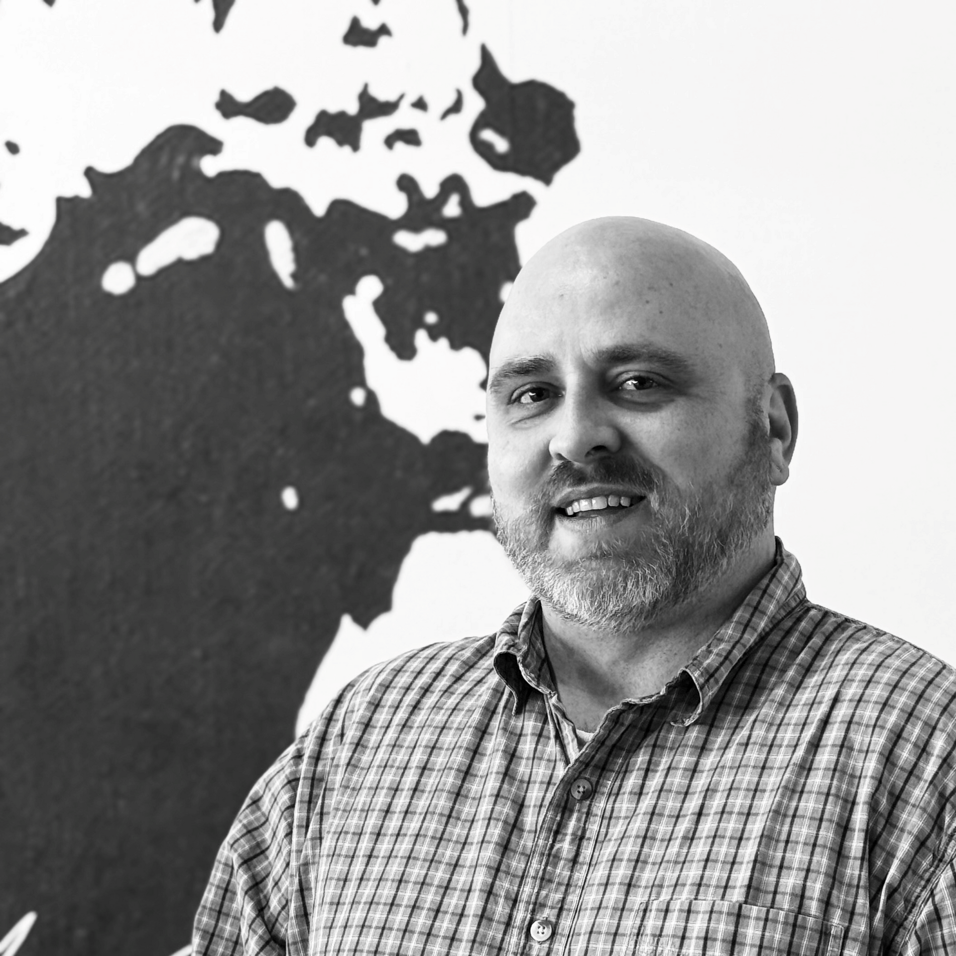 Bald man with beard smiles, wearing plaid shirt. A black and white abstract shape is in background.