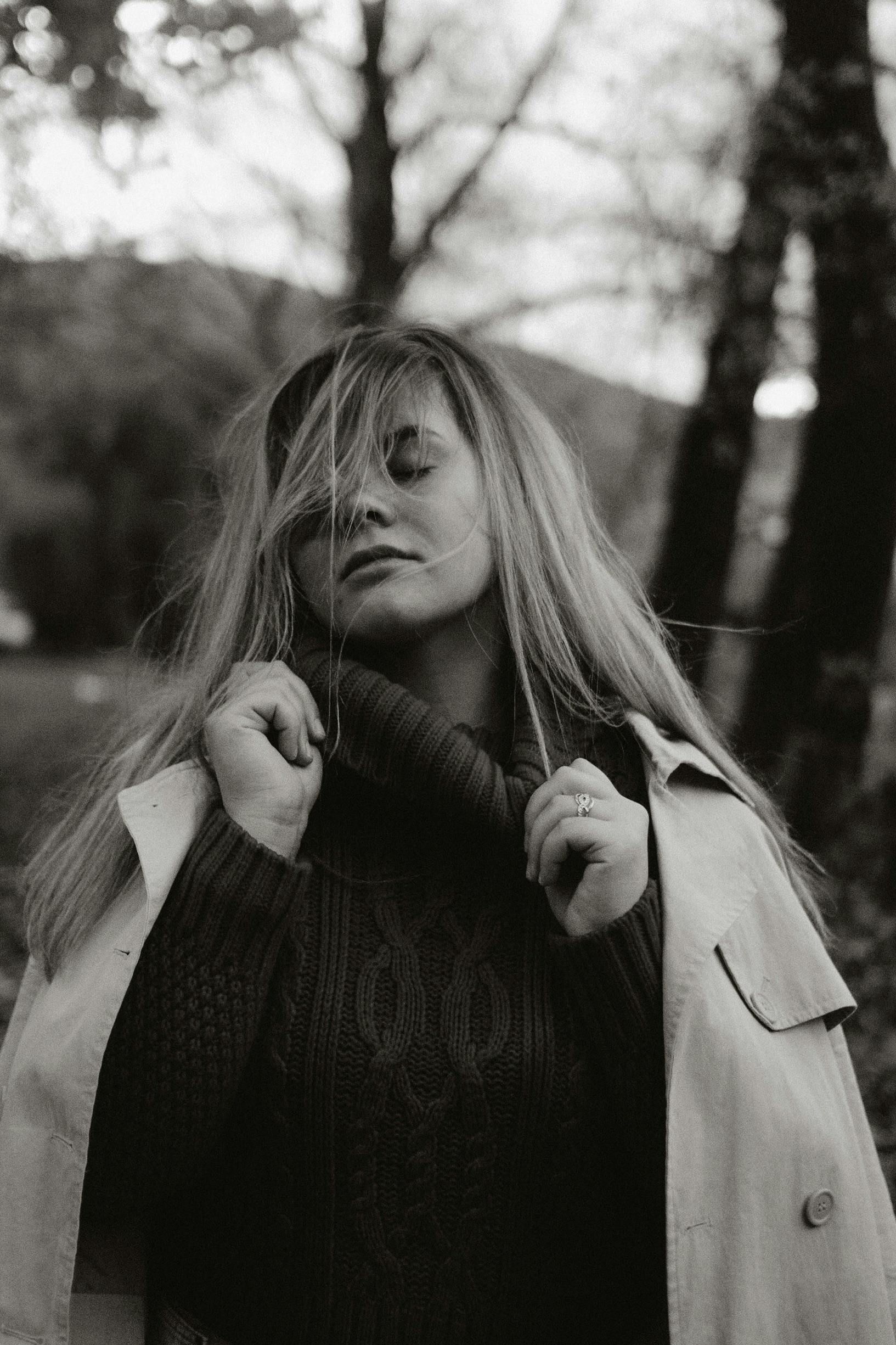 Woman with eyes closed, holding her collar in a trench coat, outside with trees.