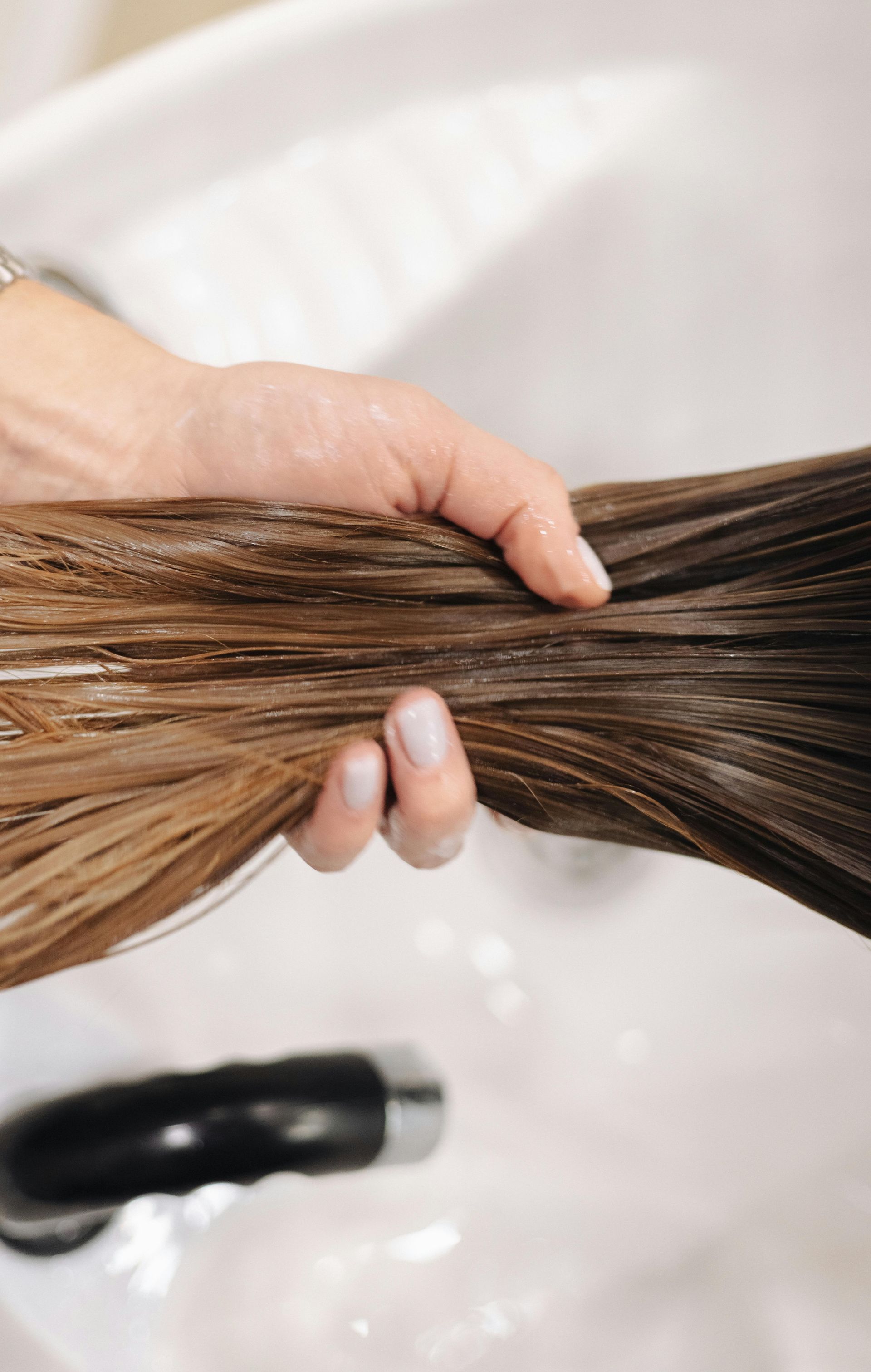 A hand holding a section of wet, brown hair over a salon sink while rinsing.