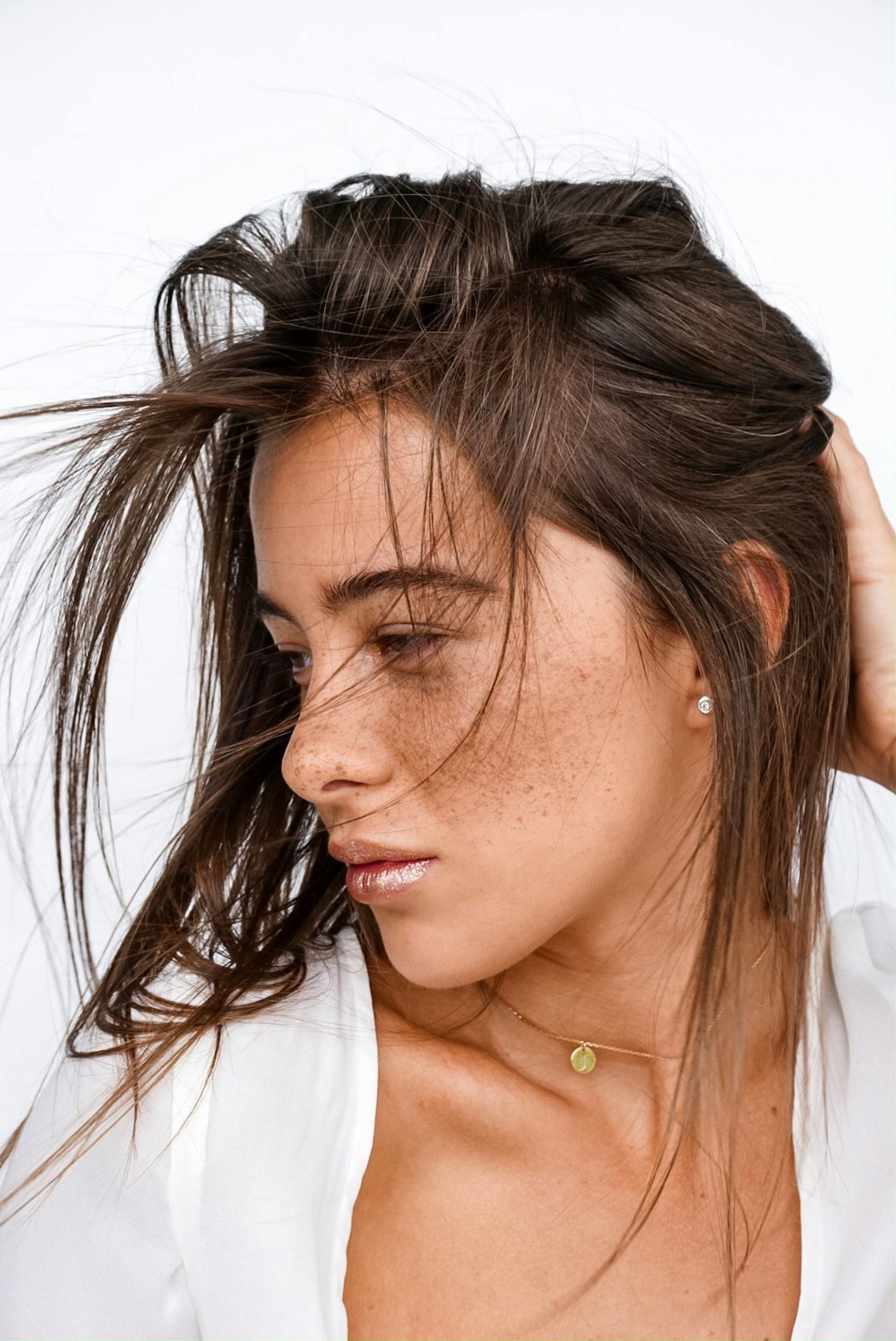 Woman with freckles and brown hair, windblown, hand in hair, wearing white top and gold necklace.