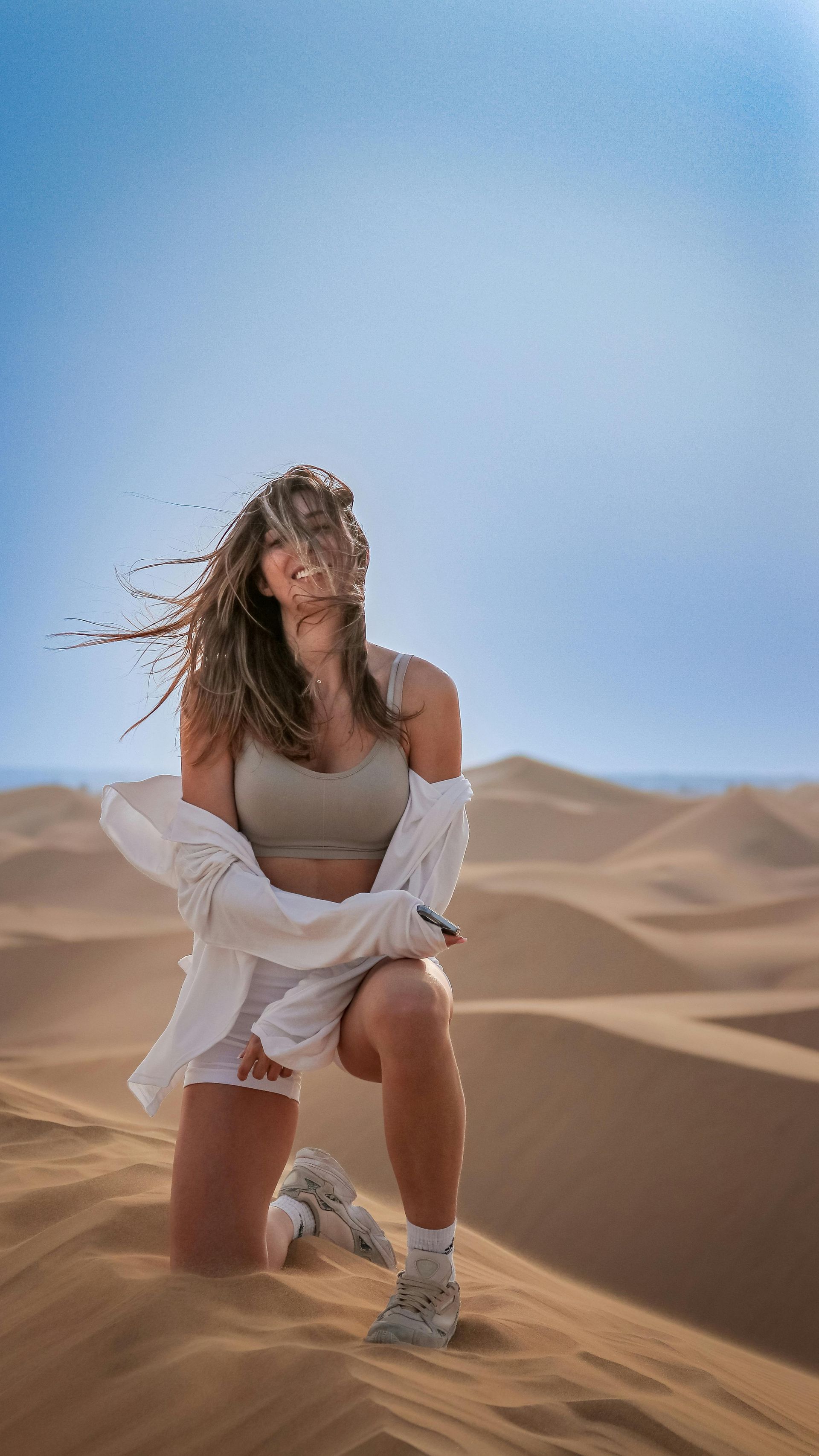 Woman kneeling in sand dunes, wind blowing hair, wearing white shirt, tan top, sneakers; bright blue sky.