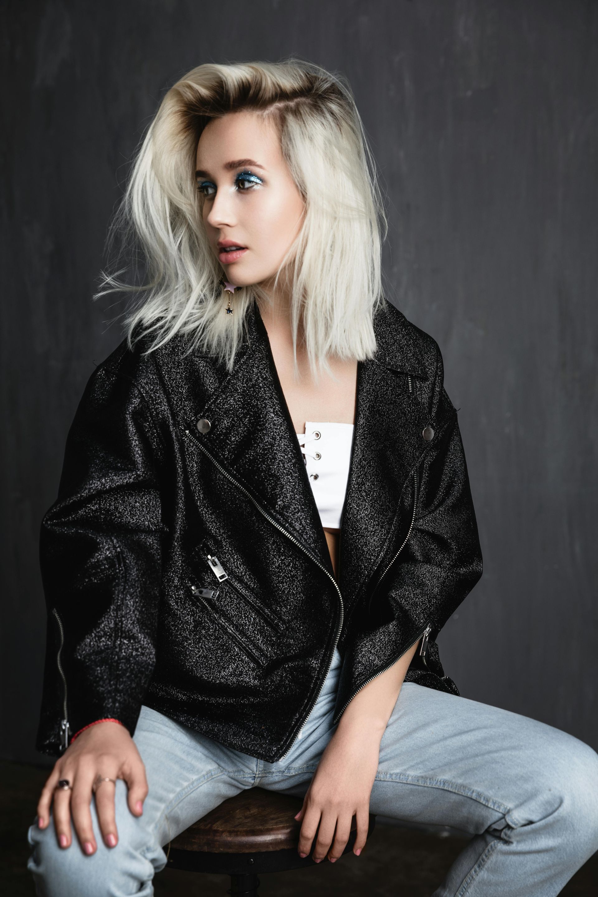 Woman with platinum blonde hair, wearing a black sparkly jacket, white top, and jeans, looking off-camera.