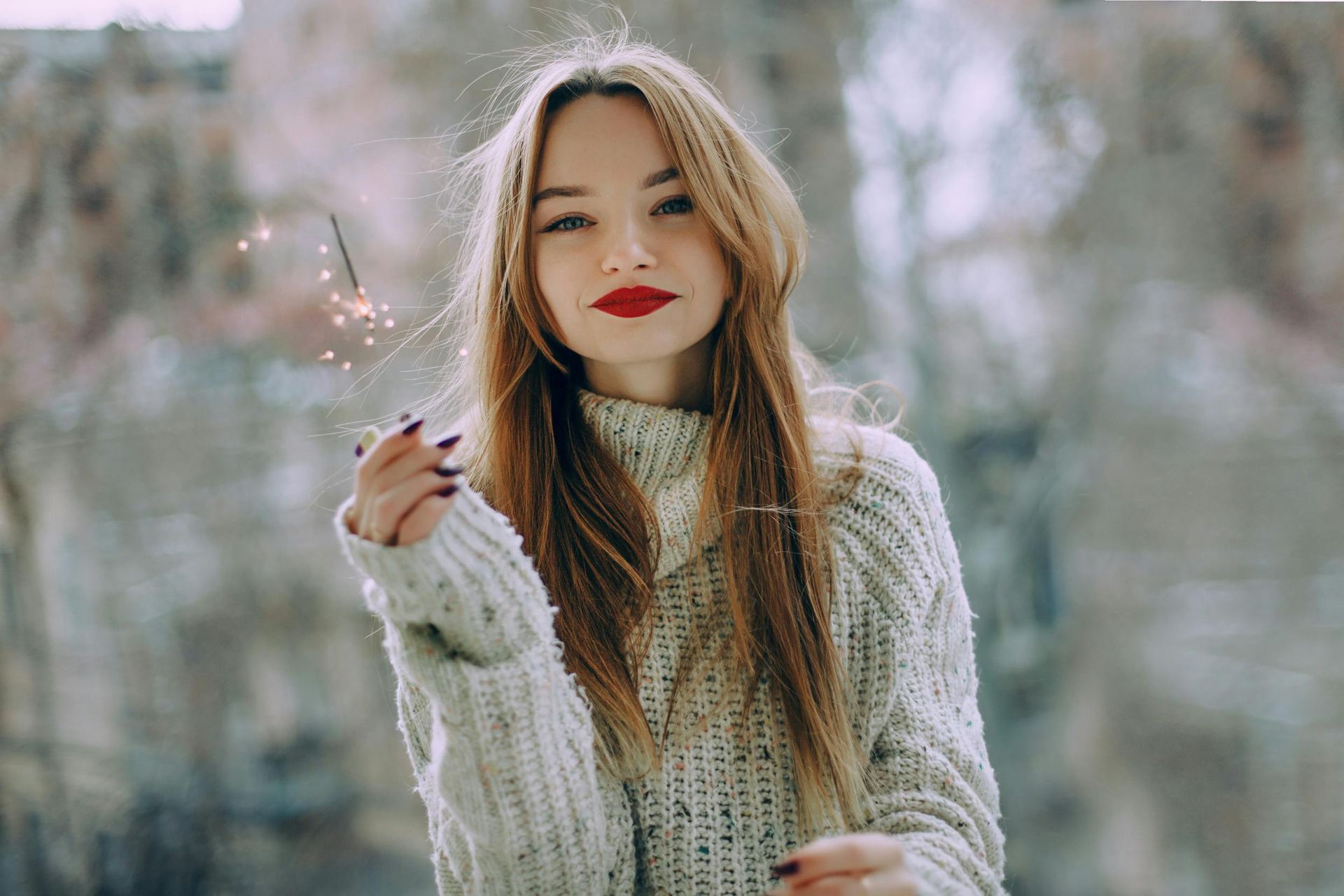 Woman with long blonde hair holds sparkler, wearing a white sweater, red lipstick, and dark nail polish.