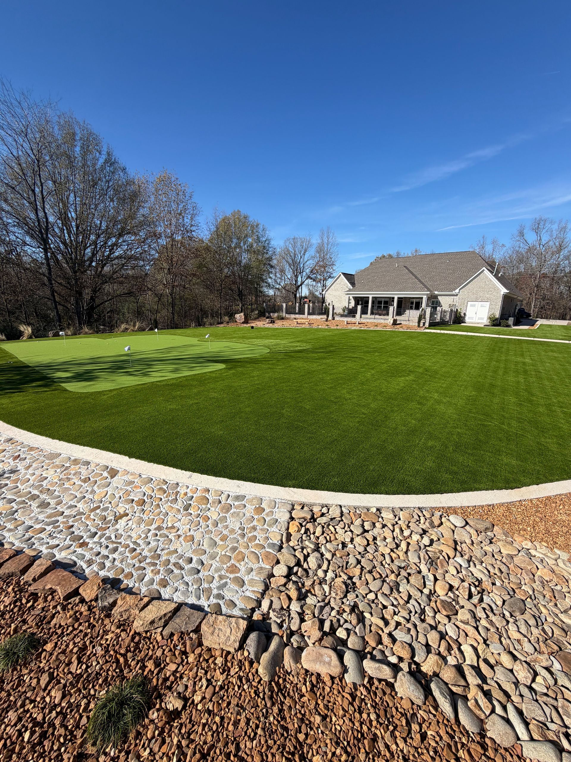 Lush green lawn curving past pebble border toward a house with a porch under a blue sky.