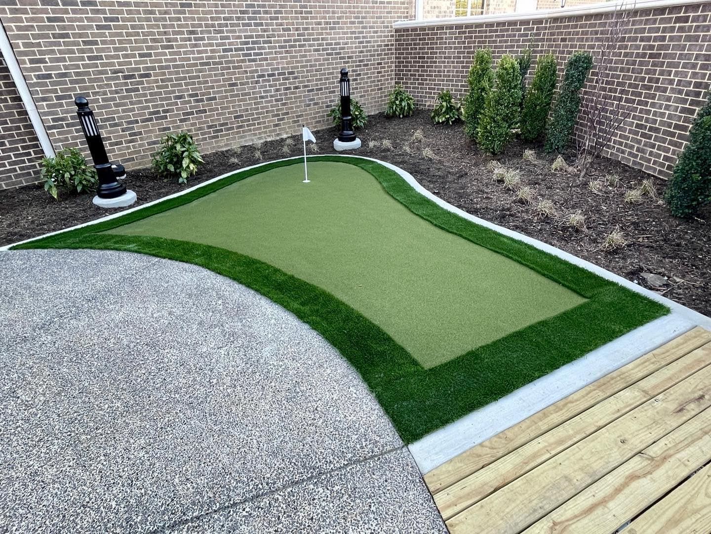 Backyard putting green with flag, surrounded by artificial turf, concrete, and brick wall.