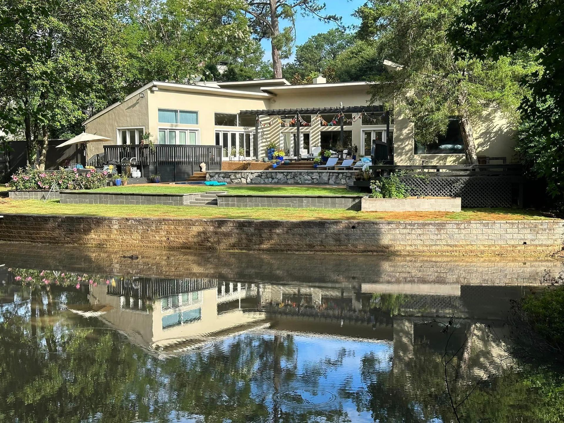 Lakeside home with flat roof, deck, and reflection in water. Trees surround.