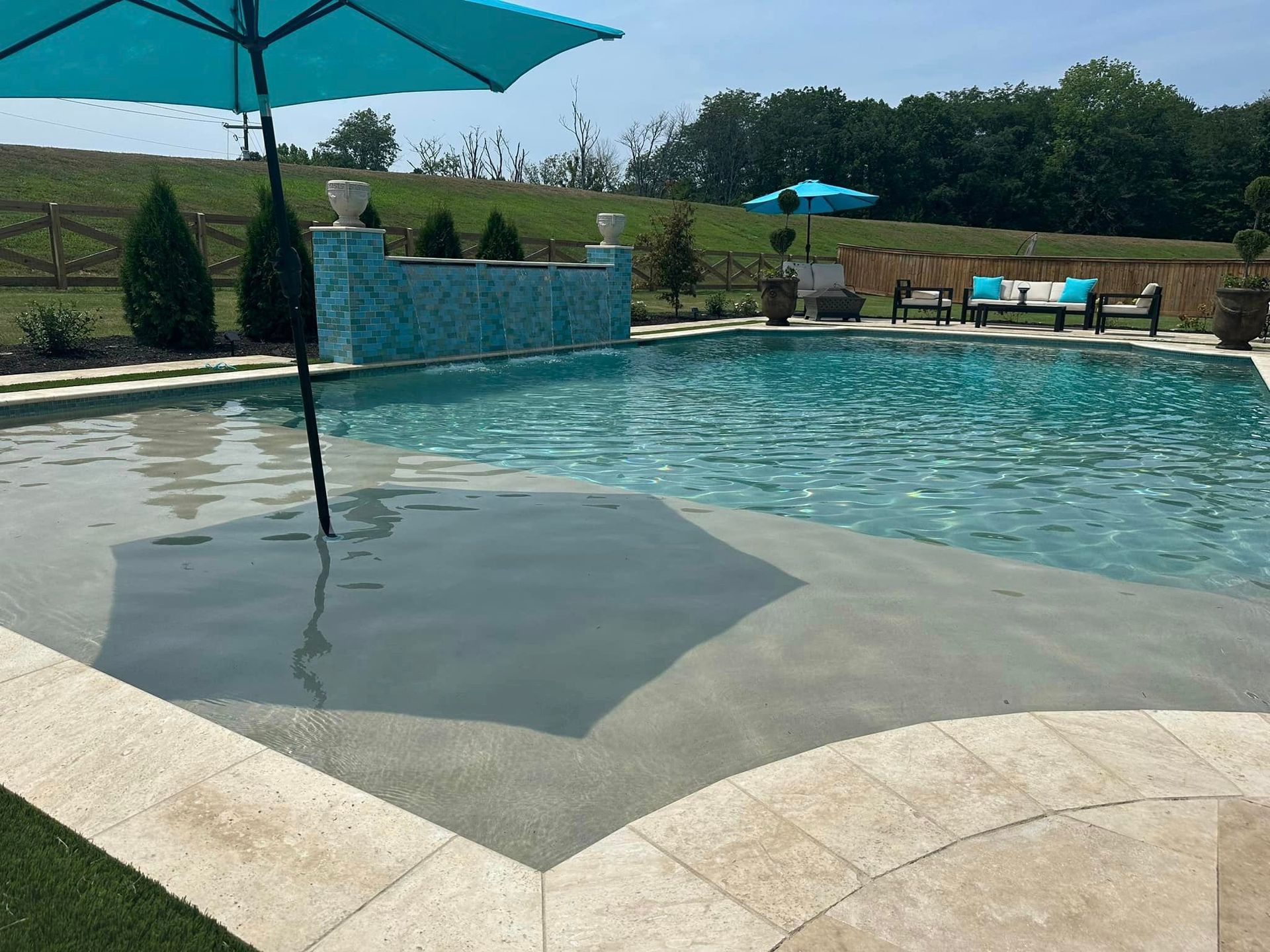 Swimming pool with turquoise water and umbrella, surrounded by stone patio, lush greenery, and a blue tile waterfall feature.