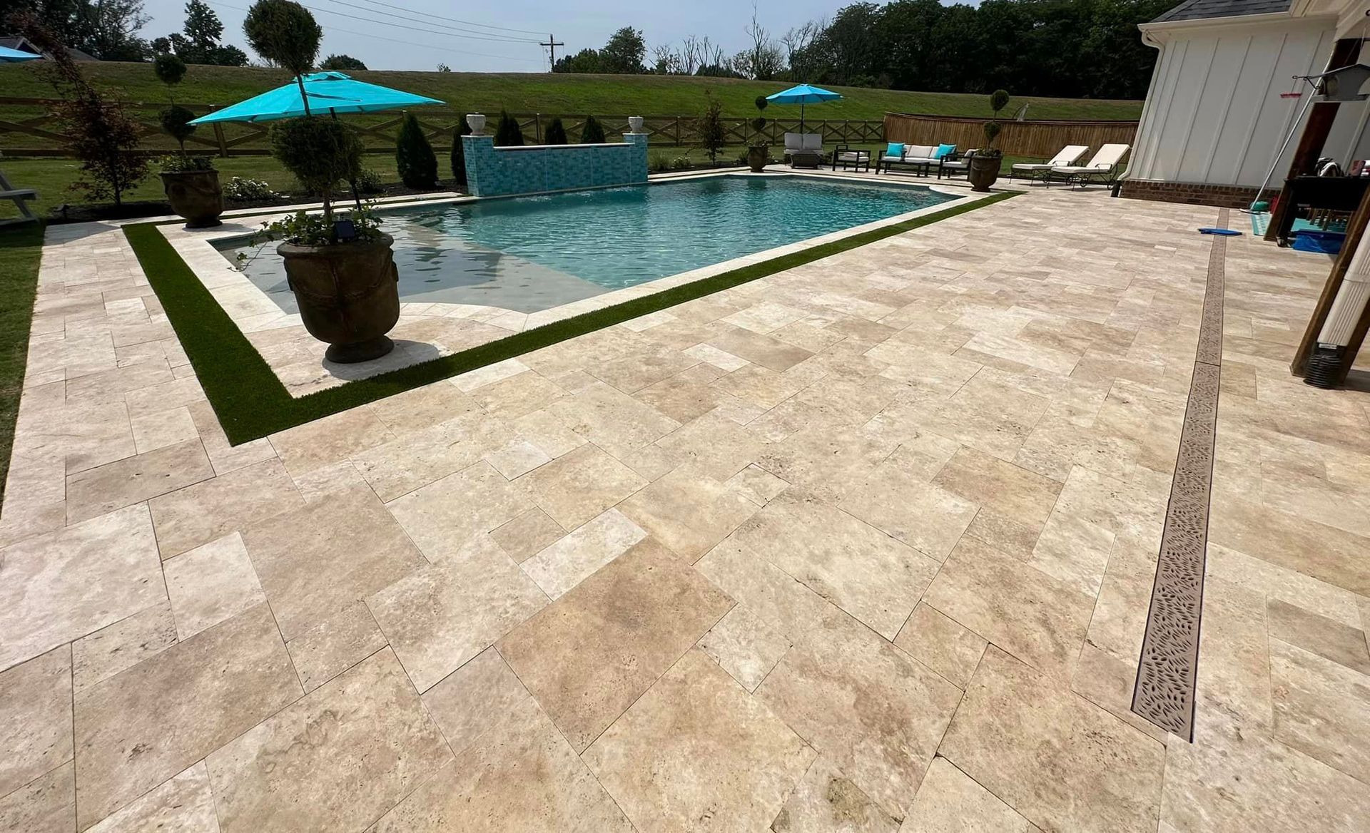 Poolside patio with travertine tiles, a pool with waterfall, and blue umbrellas.