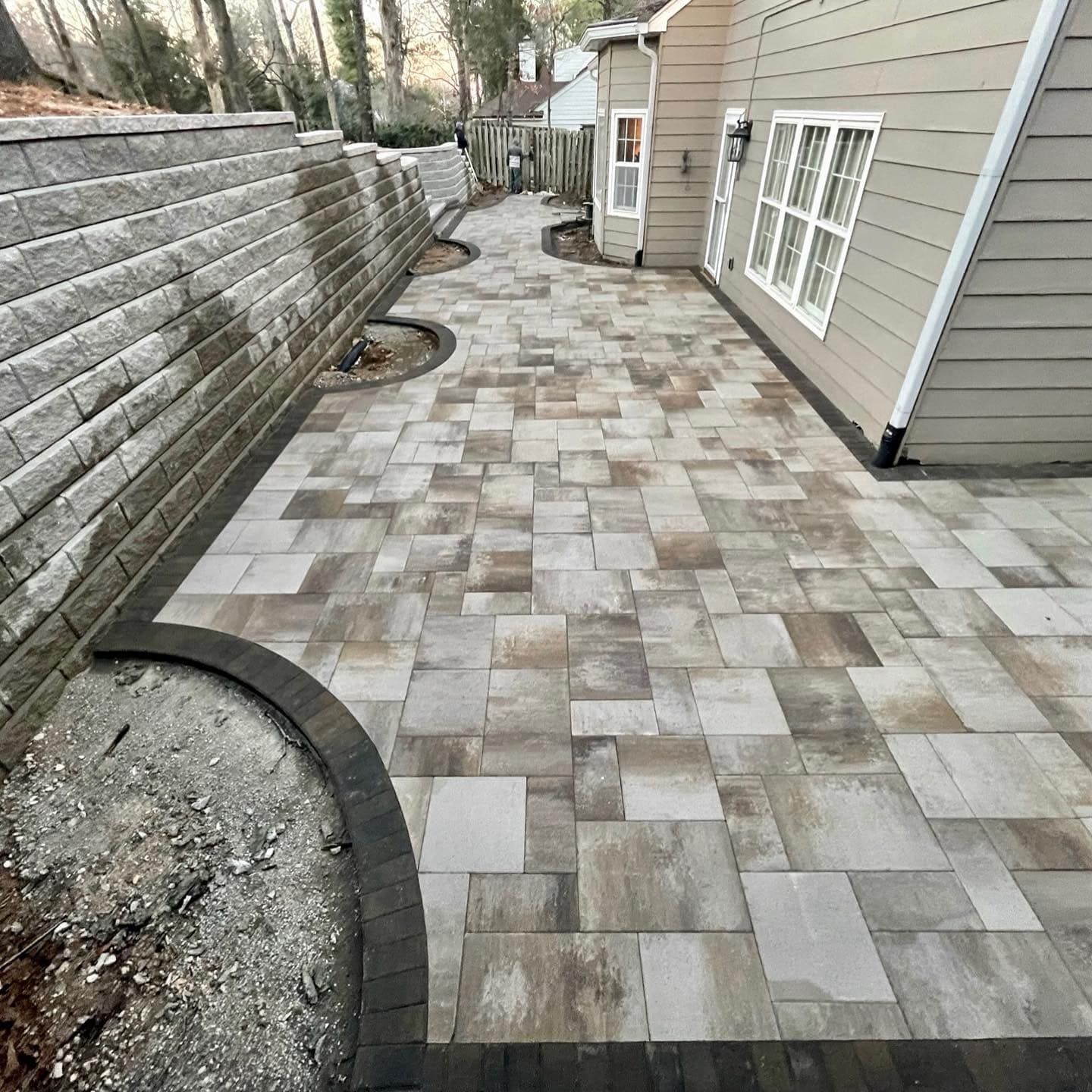 Paver patio with retaining wall and house exterior. Beige and brown pavers, dark border, and light-colored siding.