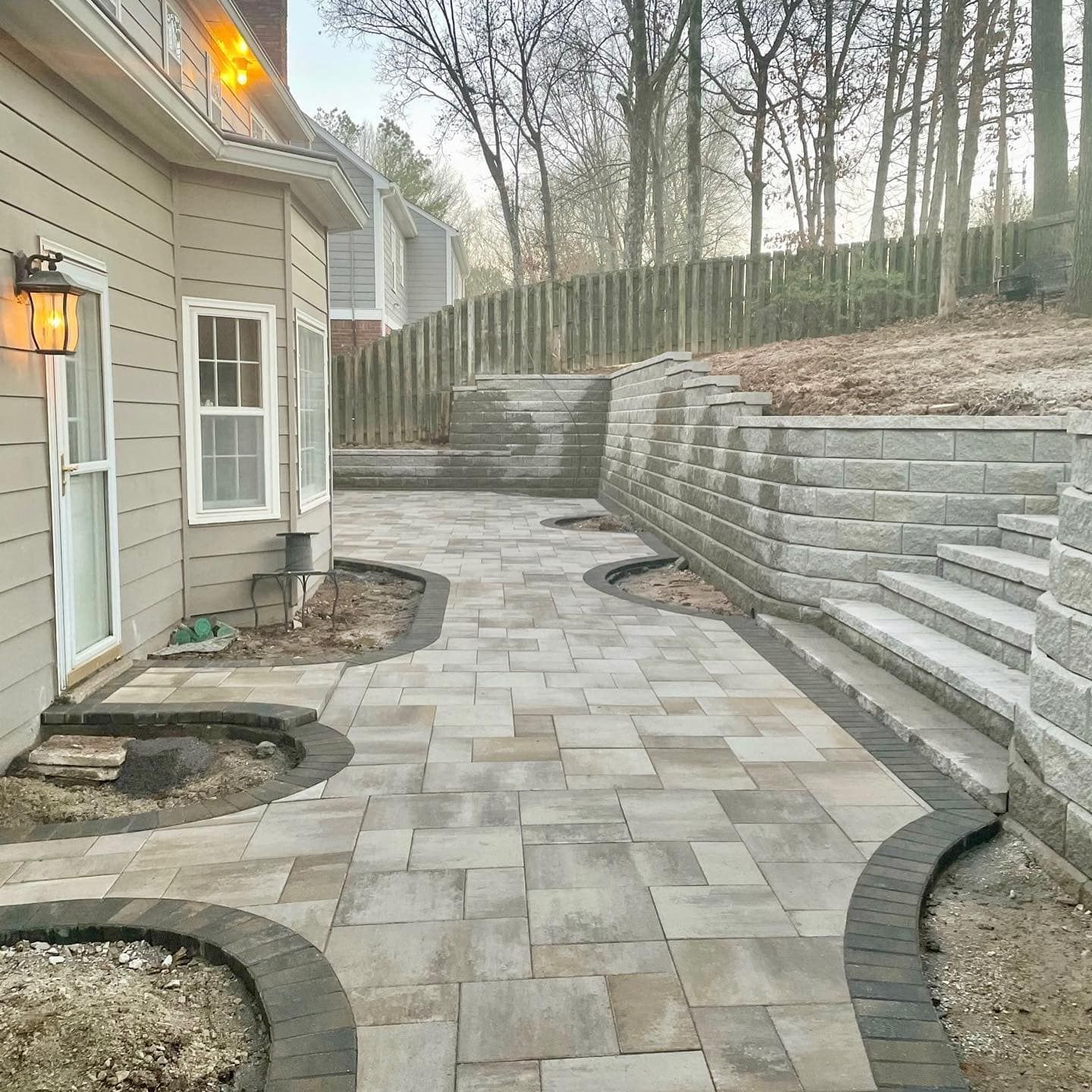 Brick patio with stone retaining walls next to a beige house, steps leading up the hillside.