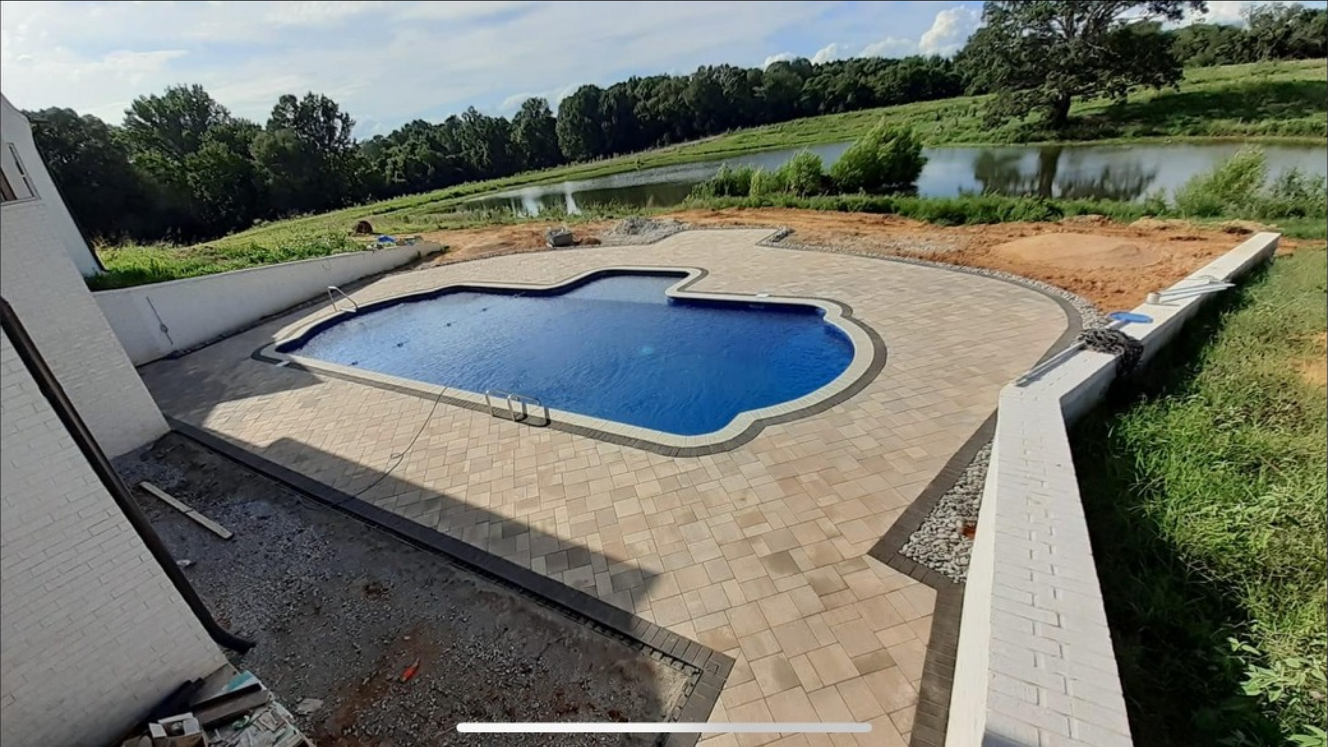 Pool with blue water surrounded by stone patio, retaining walls, and grassy hill in background.
