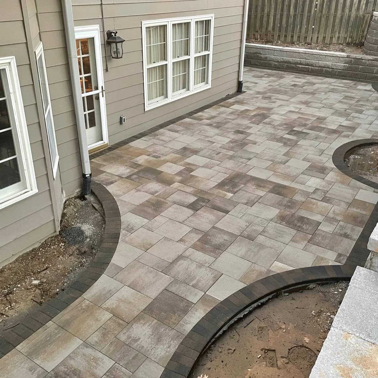A backyard patio paved with square stones in shades of gray and brown, with a curved border.