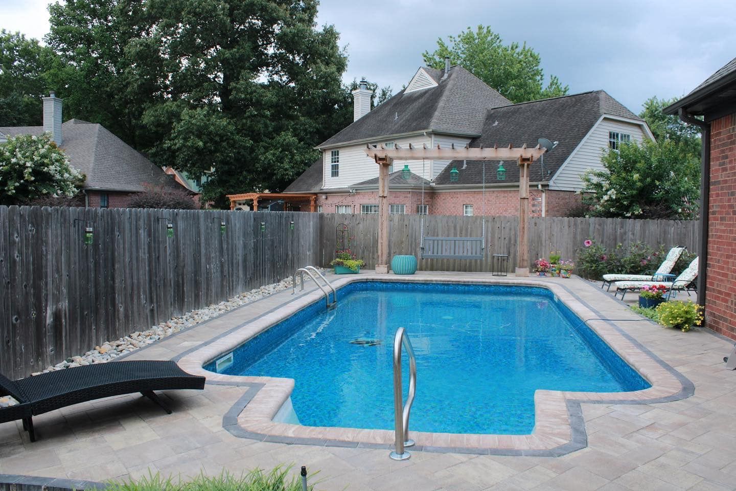 Rectangular pool in a backyard, surrounded by a concrete patio and a wooden fence, with a pergola and houses in the background.