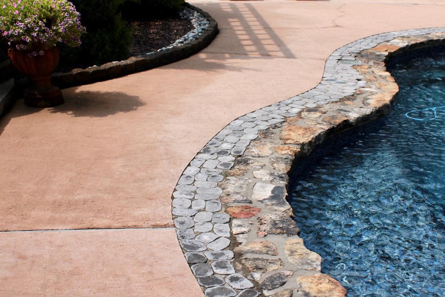 Poolside view: a curved stone border separates the blue water from brown concrete and a garden bed with flowers.