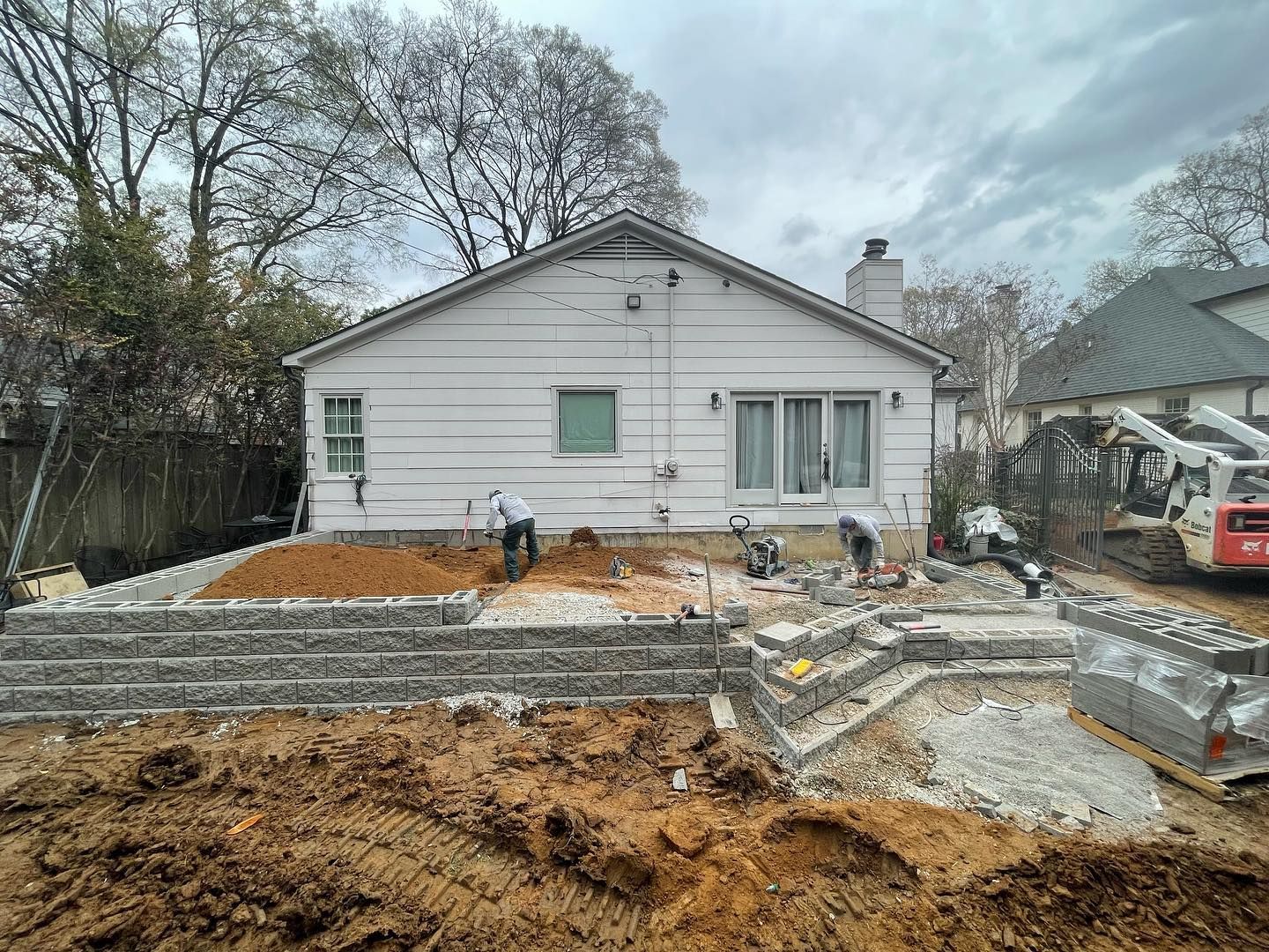 Backyard home under construction with retaining wall and workers. Cloudy sky.