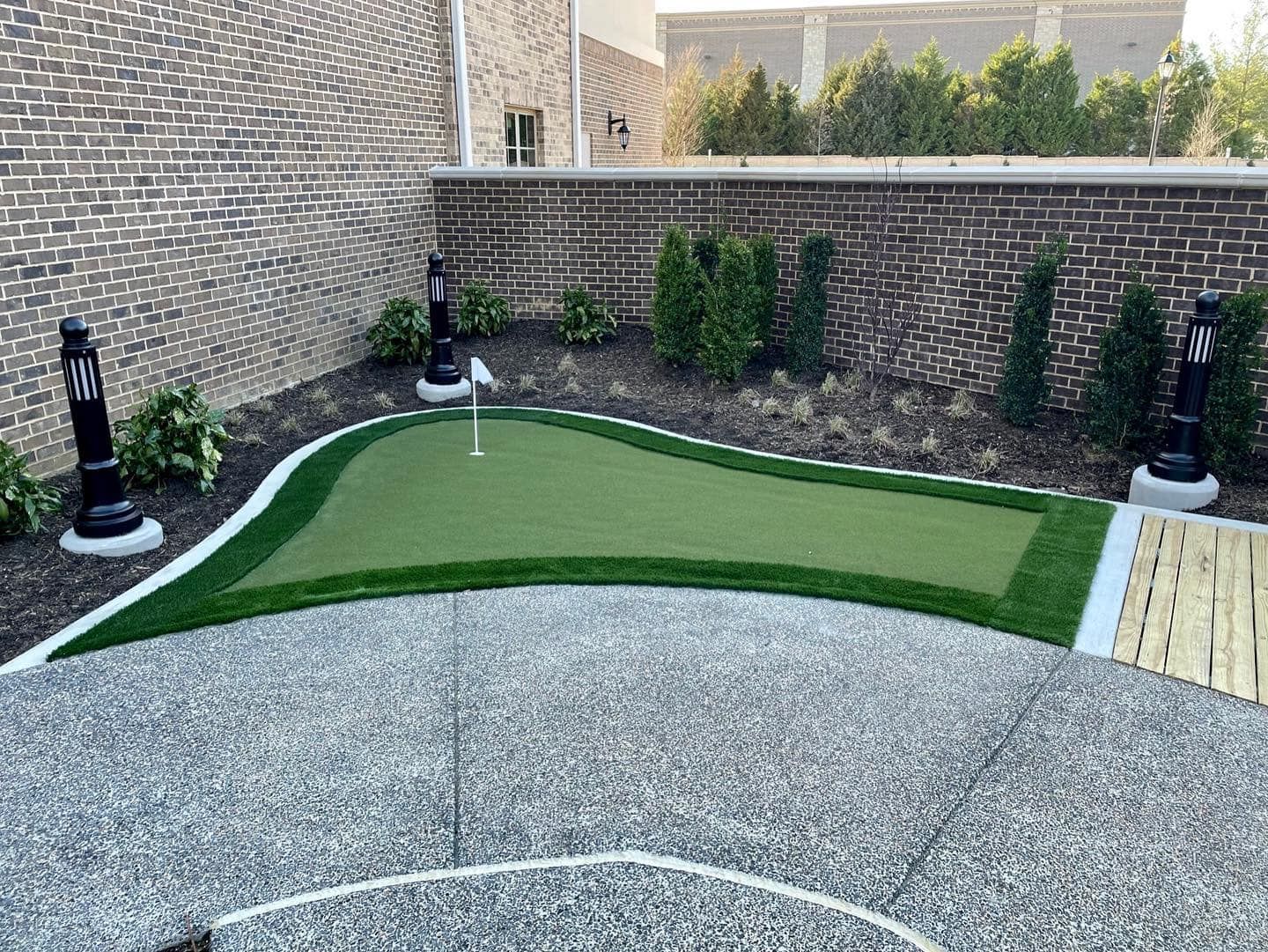 Putting green in a brick-walled courtyard, bordered by artificial turf and landscaping; flag in the hole.