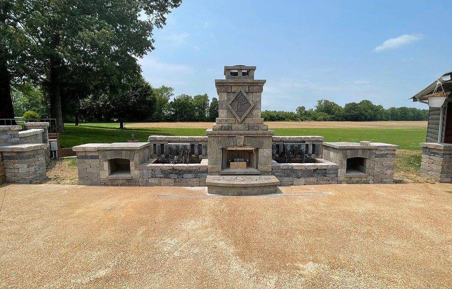 Stone outdoor fireplace with side walls, set in a yard. Blue sky and trees in the background.