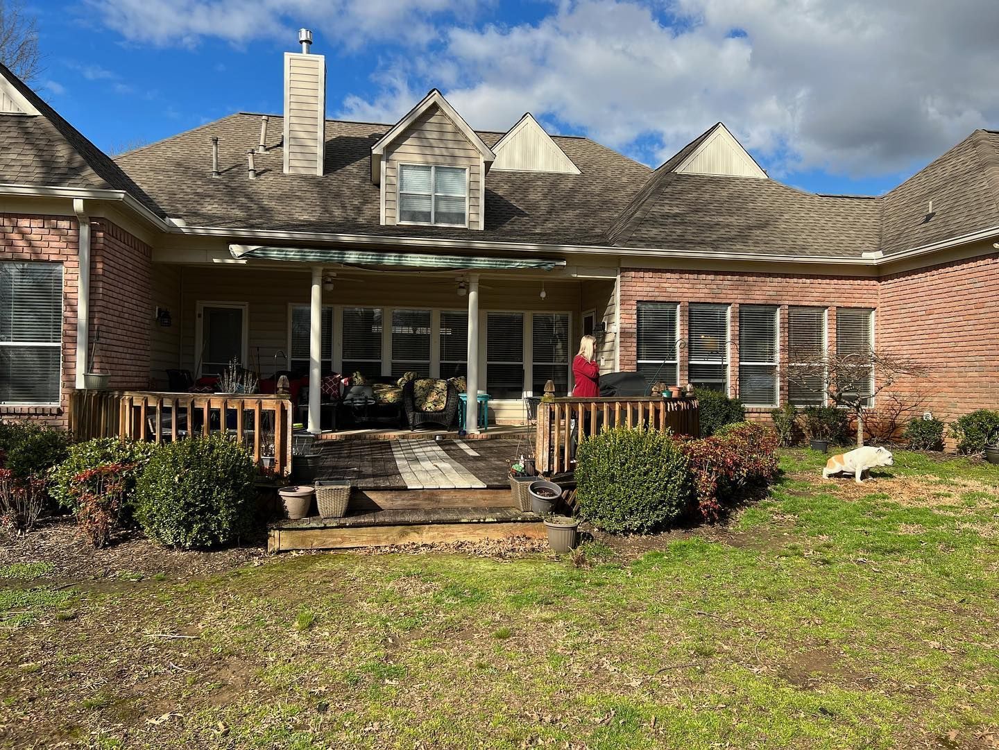 Backyard with deck, covered patio, red brick house, person on the deck, dog on grass.