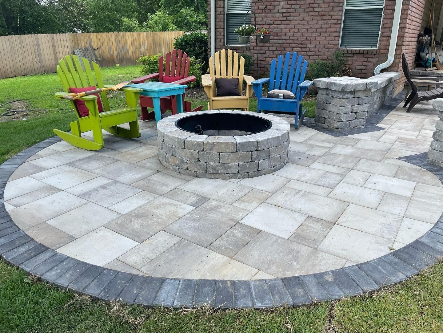 Circular patio with fire pit, colorful Adirondack chairs, brick house in background.