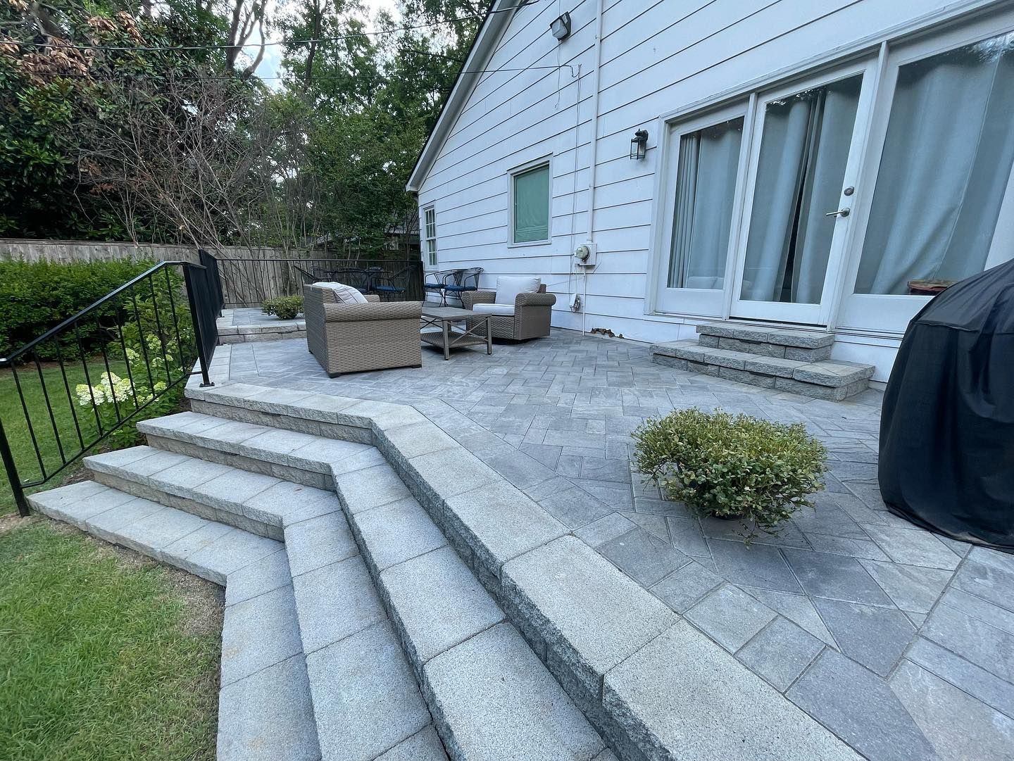Stone patio with steps leading to a yard, outdoor seating, and a grill next to a white house.