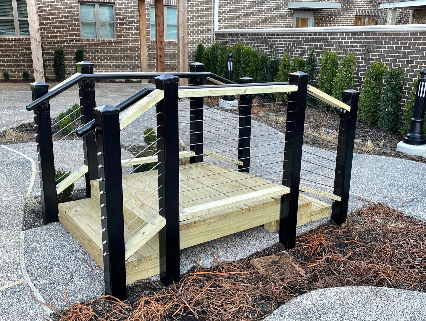 Wooden platform with black handrails and steps. Outdoor setting with gravel and brick building.