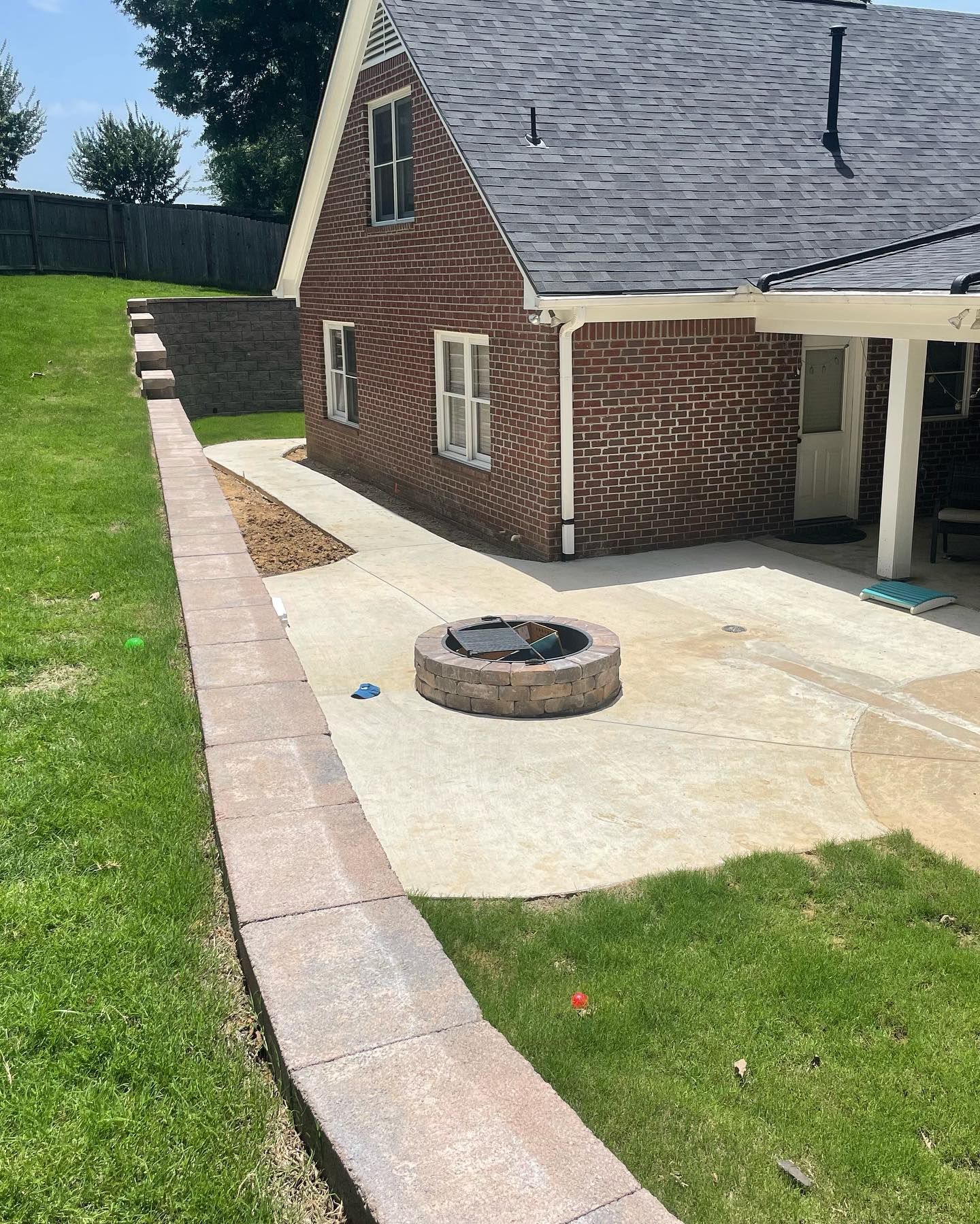Brick house with retaining wall, patio, and fire pit. Green grass and blue sky are visible.