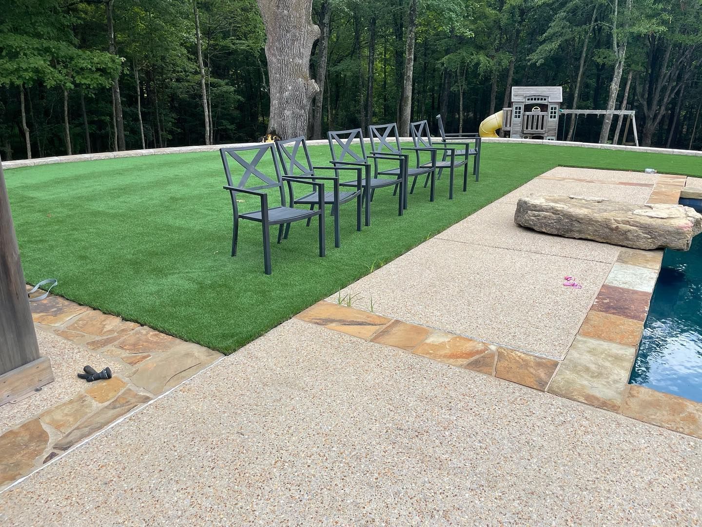 Lawn area with chairs, a pool, and stone patio. Trees in background.
