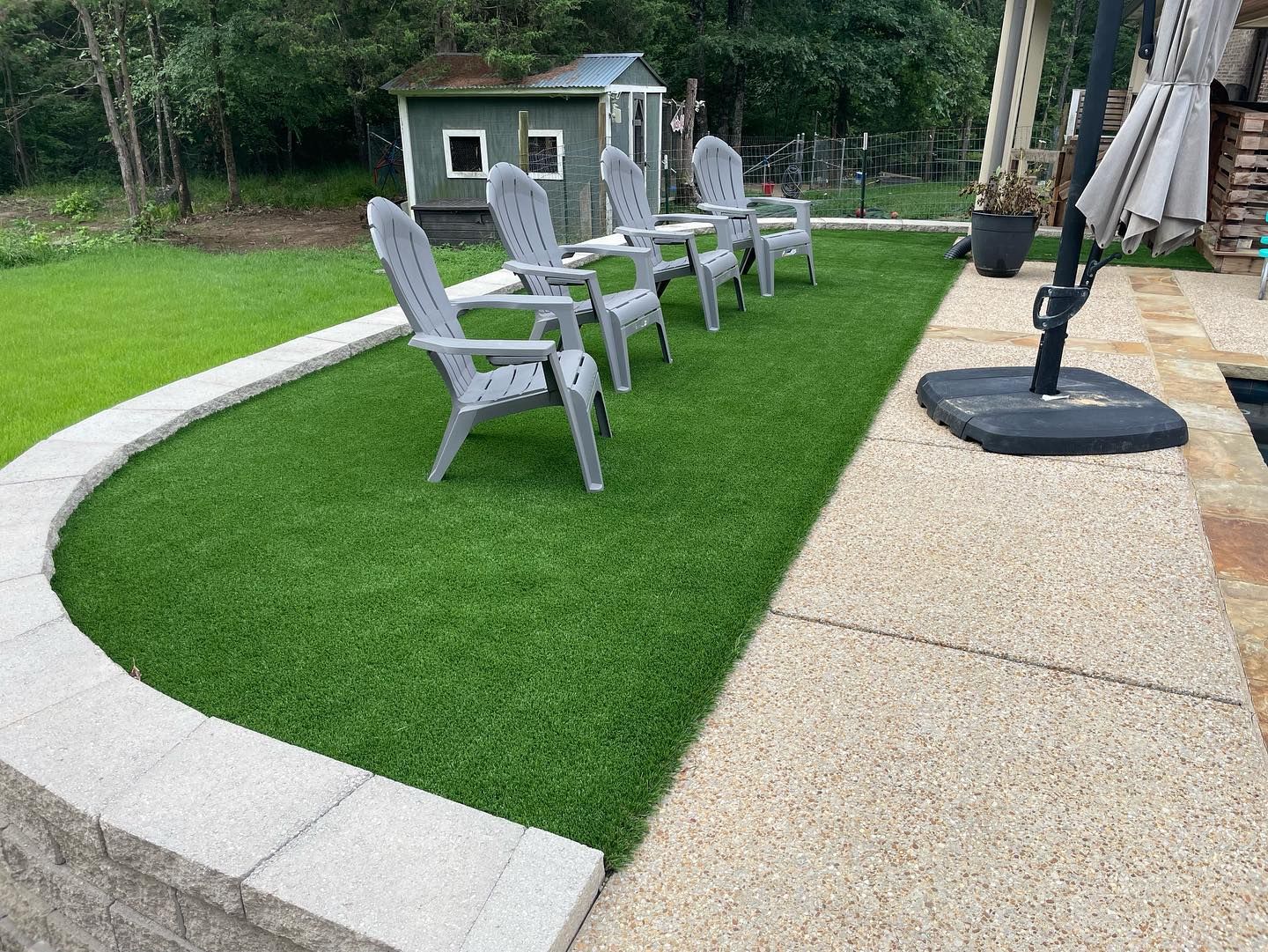 Lush green artificial turf area with gray chairs, next to a stone patio and retaining wall.