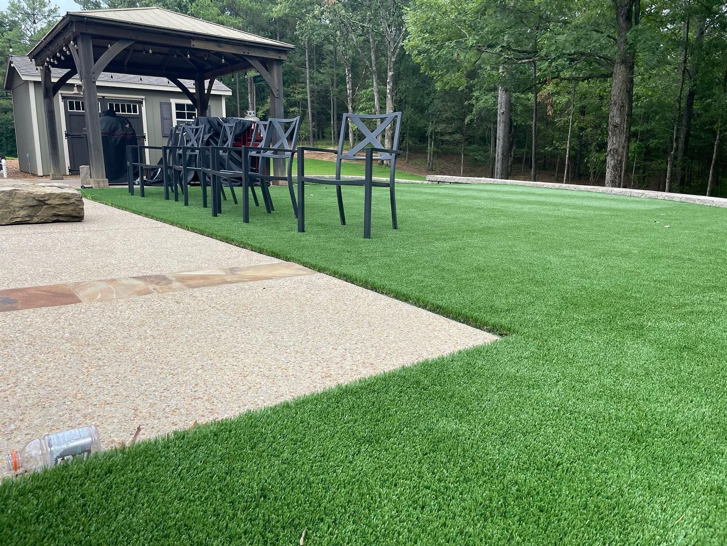 Artificial turf lawn with gravel patio, black chairs under a pergola, and small shed in the background.