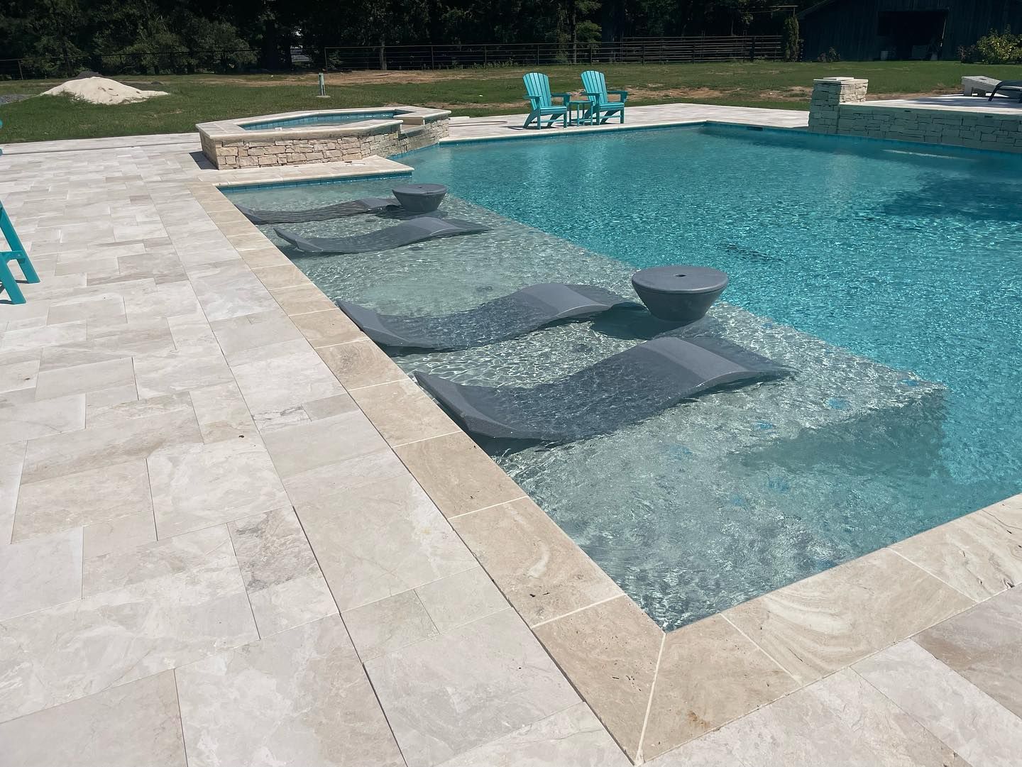 Pool with in-water lounge chairs and a small table. Turquoise water, light stone patio, and two chairs in the background.