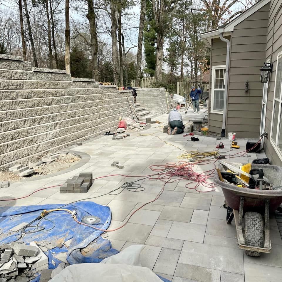 Workers constructing a stone patio and retaining wall near a house. Tools and materials are scattered around.