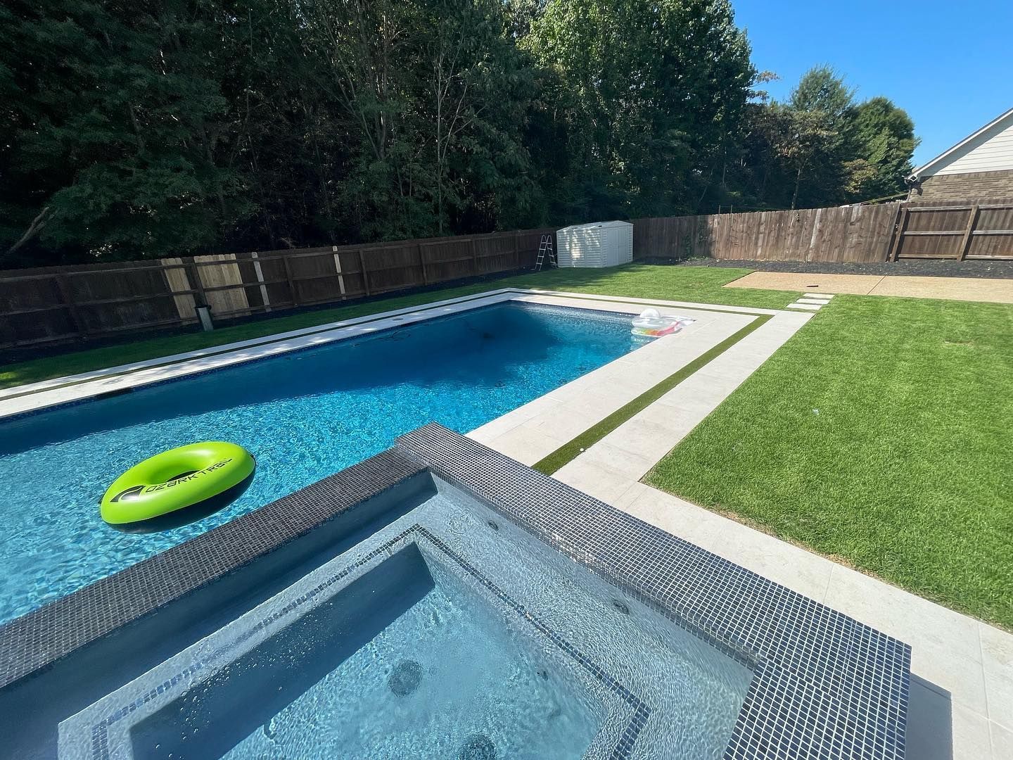 Backyard pool with spa, grass, and a wooden fence under a blue sky. A green inflatable floats in the pool.