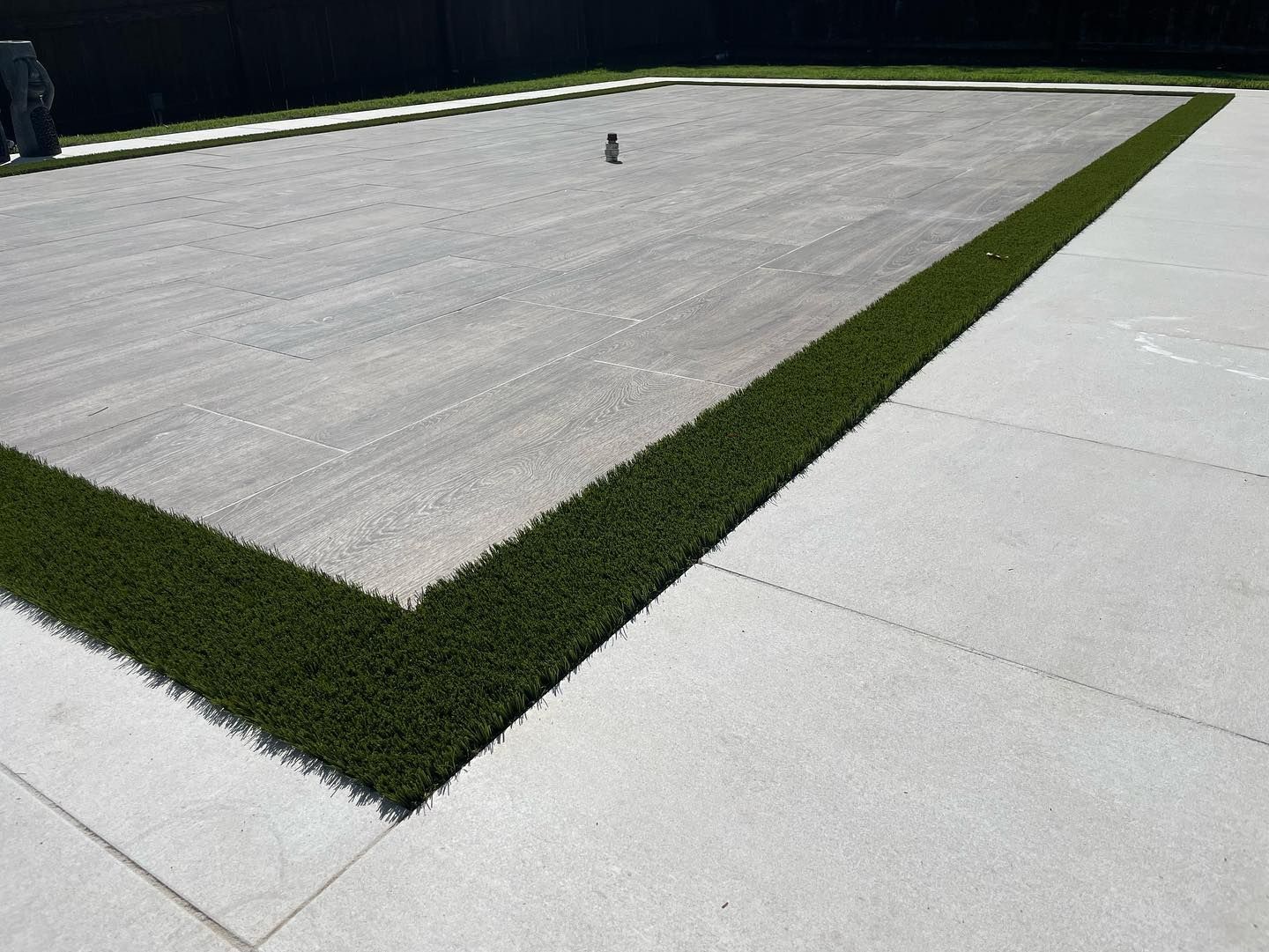 Rectangle of gray base material framed with dark green artificial grass, set on a light gray concrete patio.