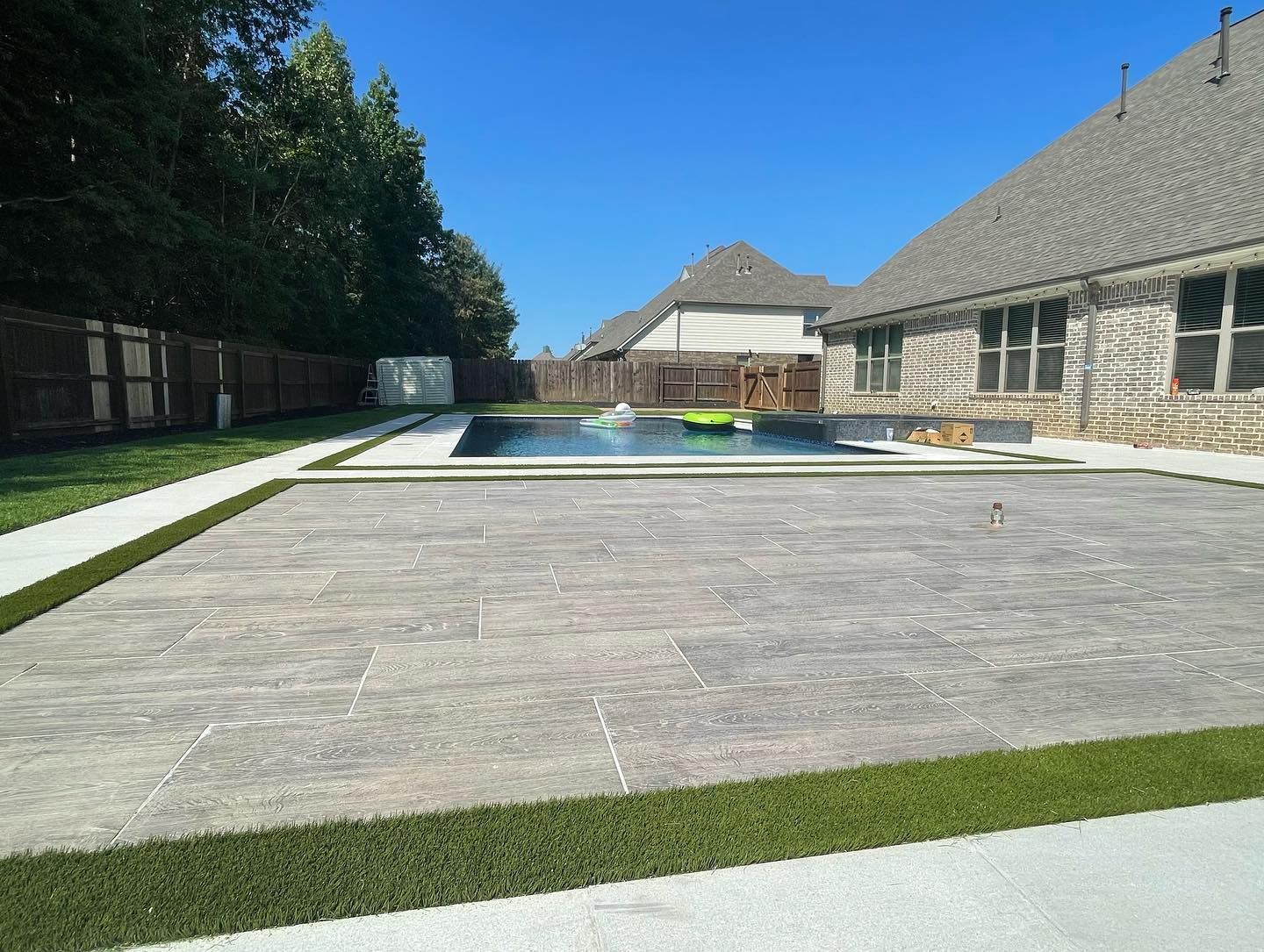 Backyard with pool, patio, house, and green turf edging under a bright blue sky.
