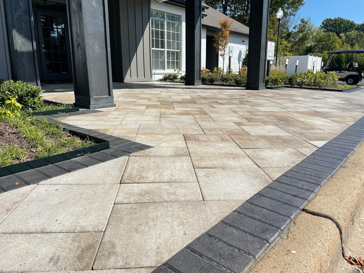 Paver patio with dark border leading to a building with black columns and a glass door.