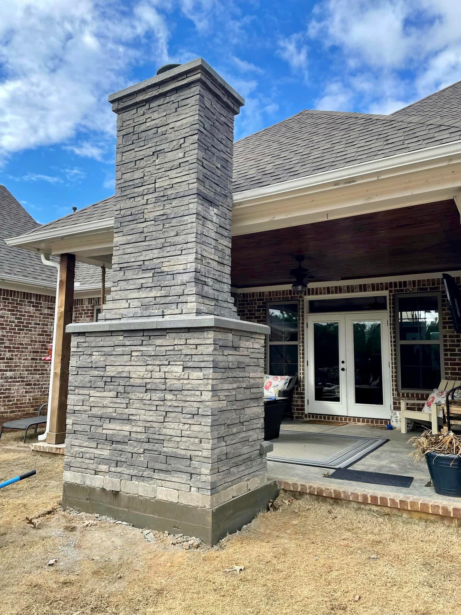 Outdoor brick chimney with gray stone facade, standing next to a house with a covered patio.