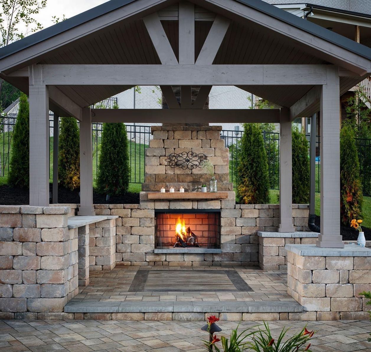 Outdoor stone fireplace under a covered patio with burning fire and surrounding stonework.