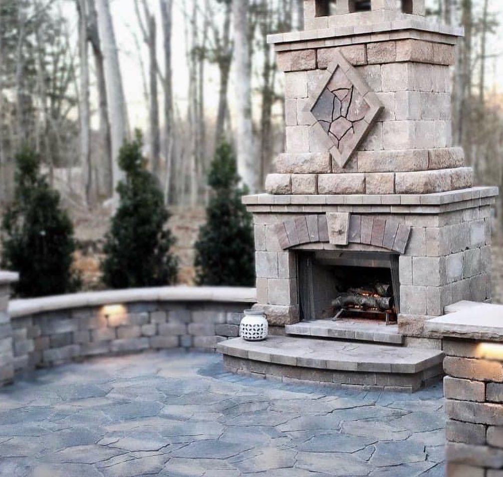 Outdoor stone fireplace on a paved patio with surrounding wall and trees in the background.