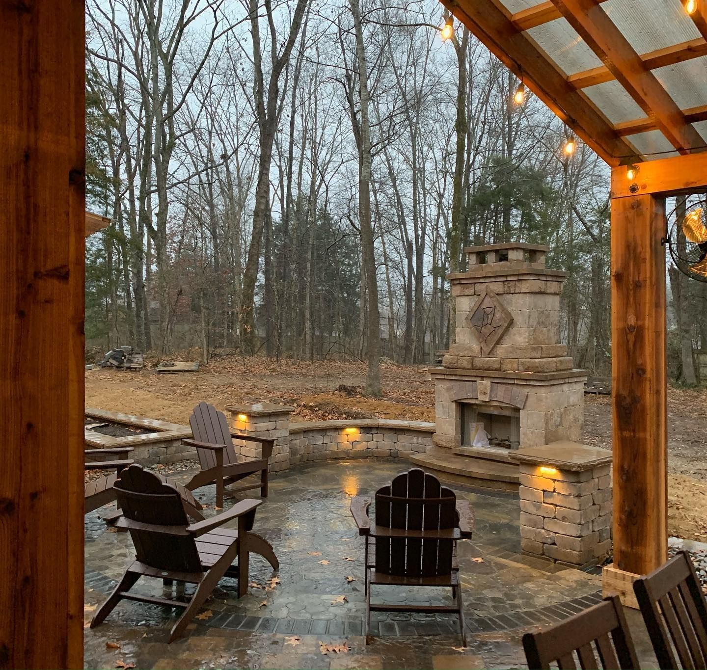 Outdoor patio with stone fireplace, wooden chairs, and string lights under a covered area in a wooded setting.