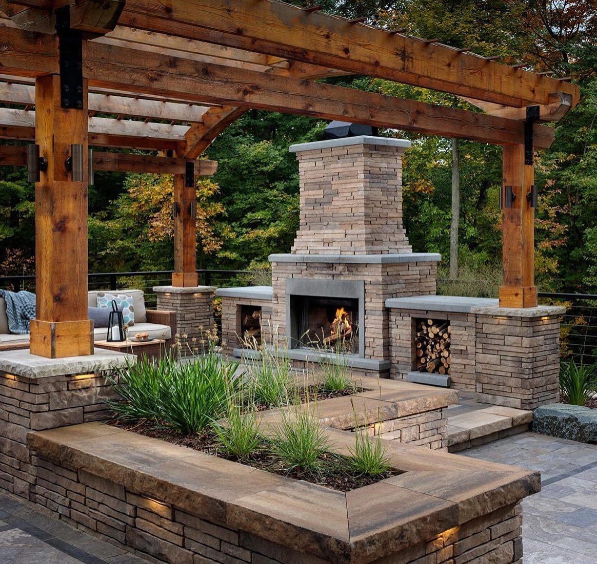 Outdoor fireplace with wooden pergola, stone structure, and built-in seating surrounded by greenery.