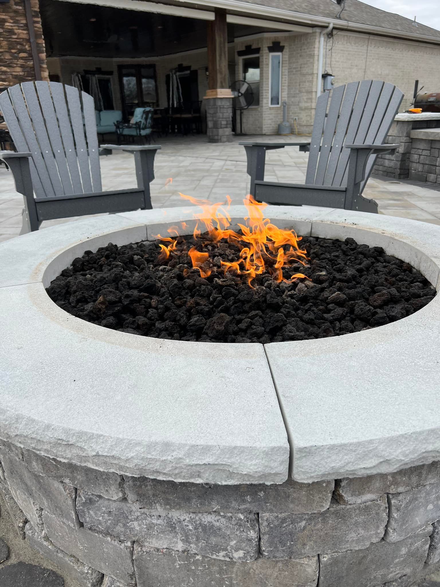 Fire pit with burning flames, two gray Adirondack chairs on patio.
