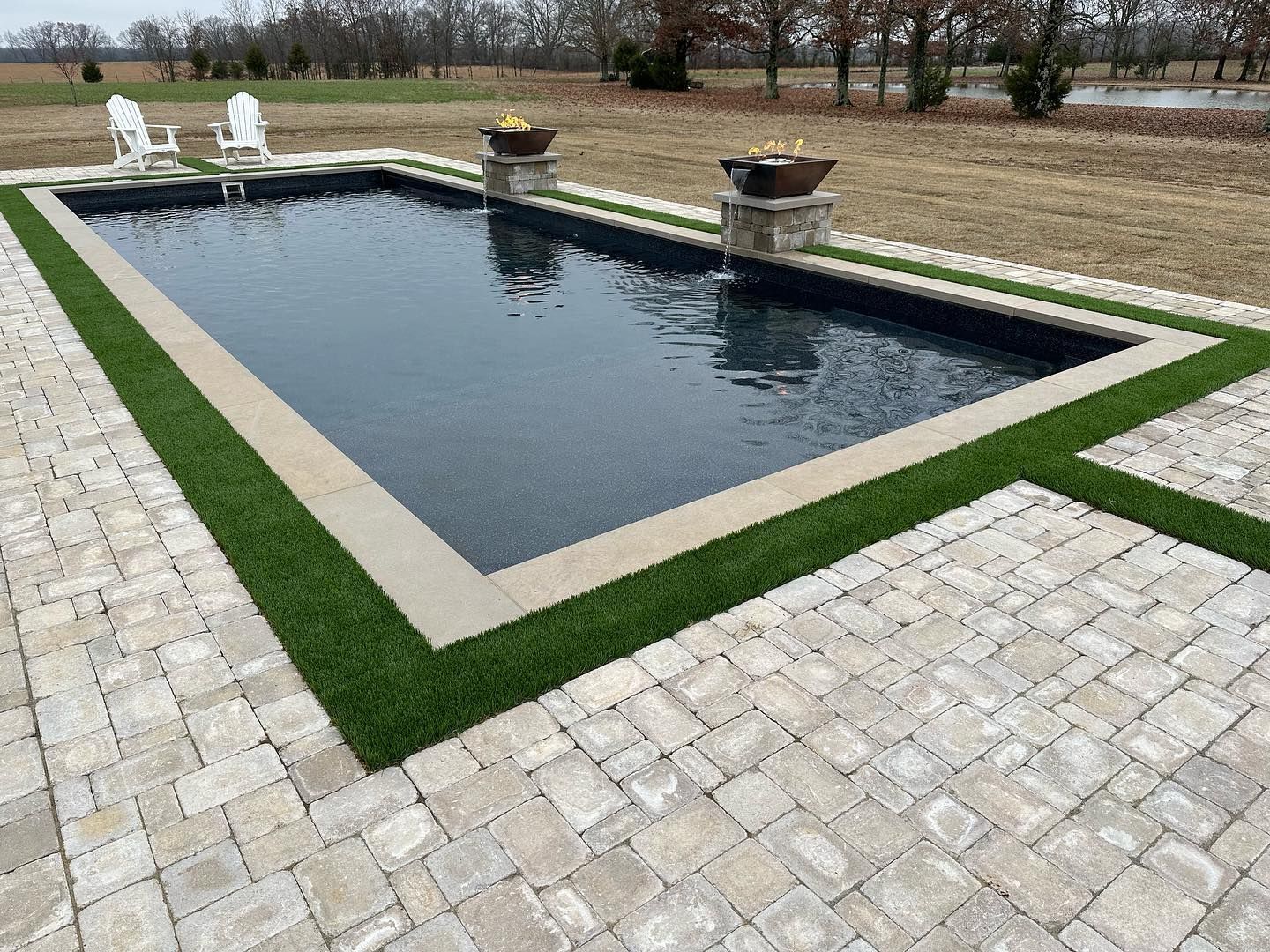 Rectangular outdoor pool with stone border, green turf, and brick patio. Two chairs and fire pits.