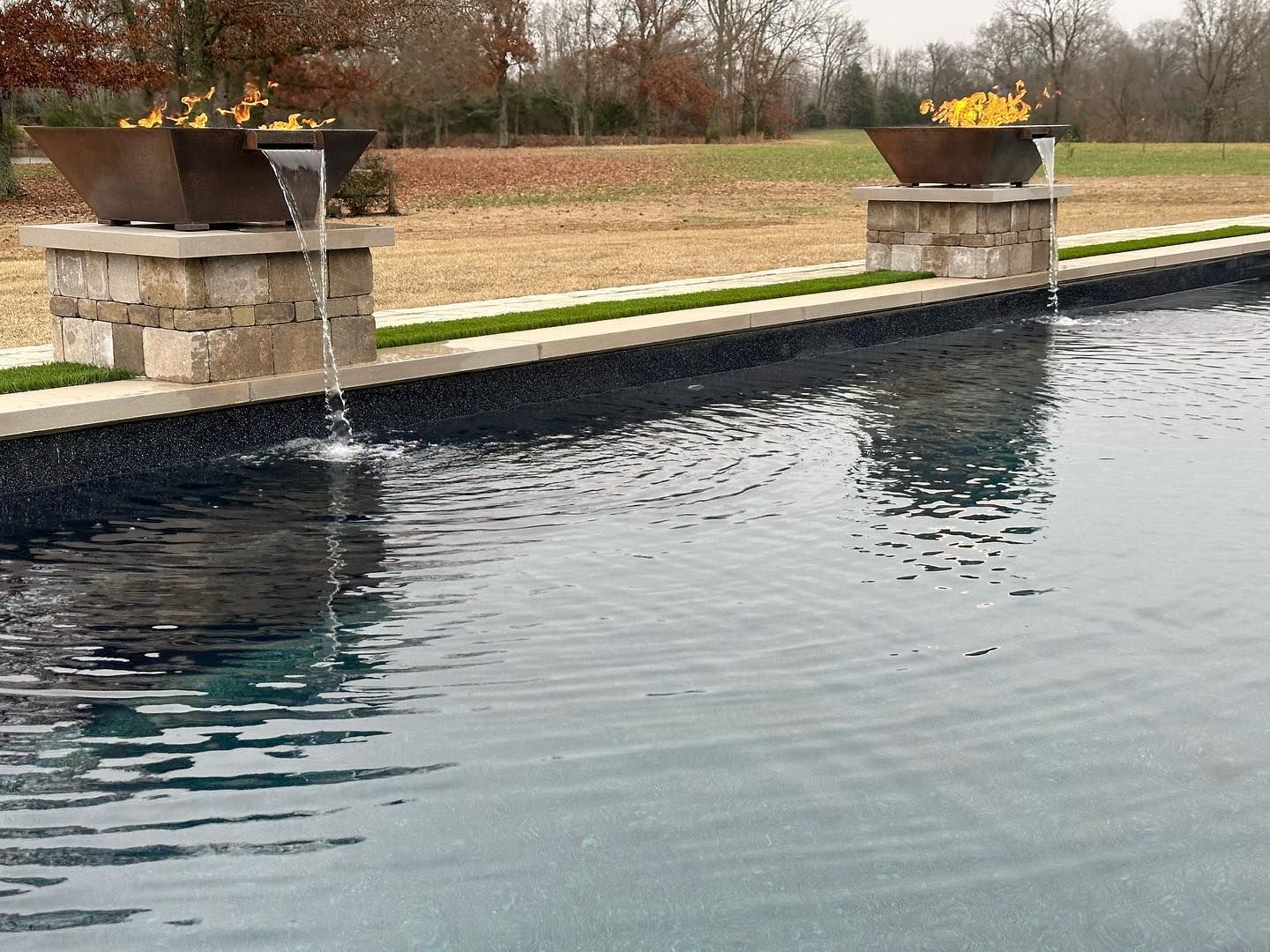 Two water features with flowers beside a pool. Water flows from metal planters atop stone columns.