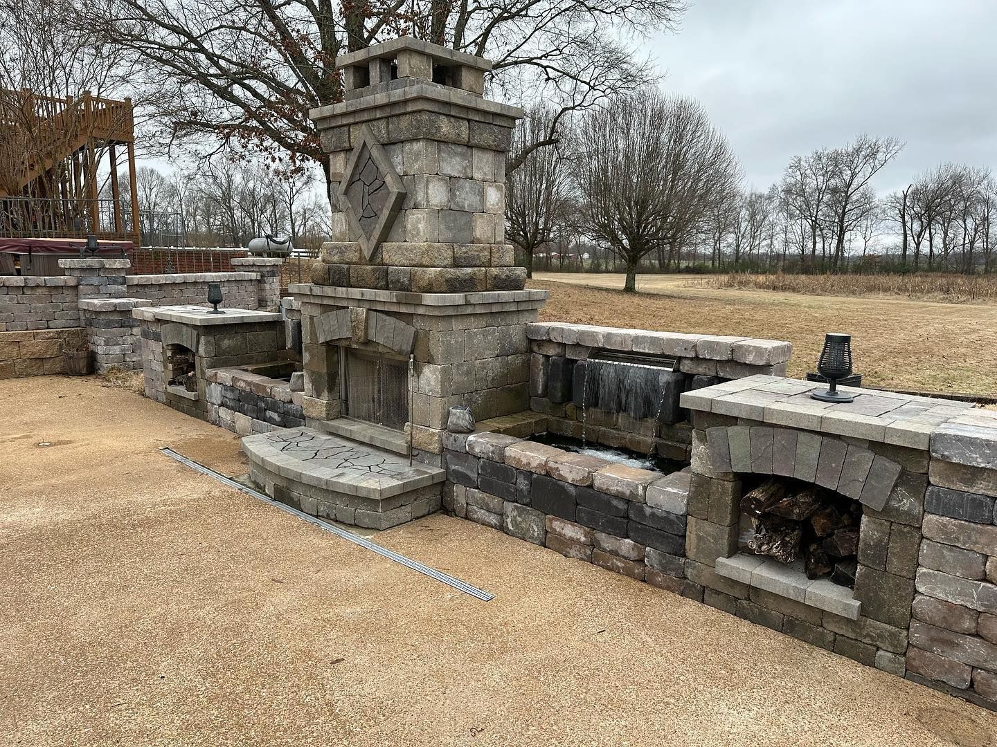 Outdoor stone fireplace with built-in features: waterfall, shelving, and chimney. Field and trees in background.