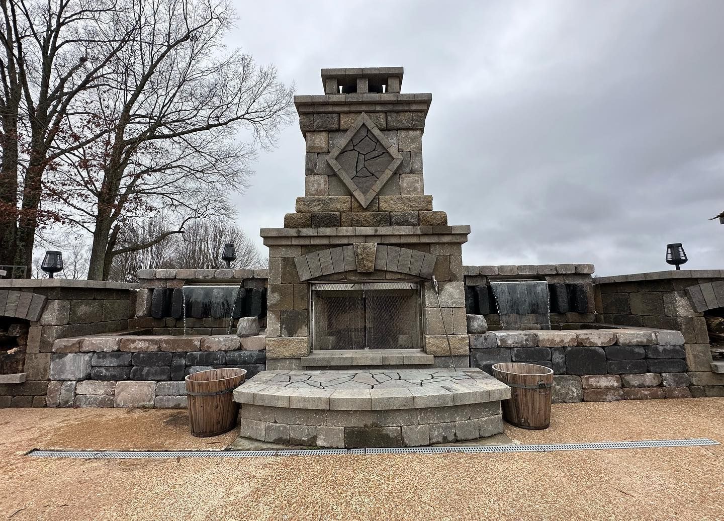Stone outdoor fireplace with diamond design, set against a water feature and cloudy sky.