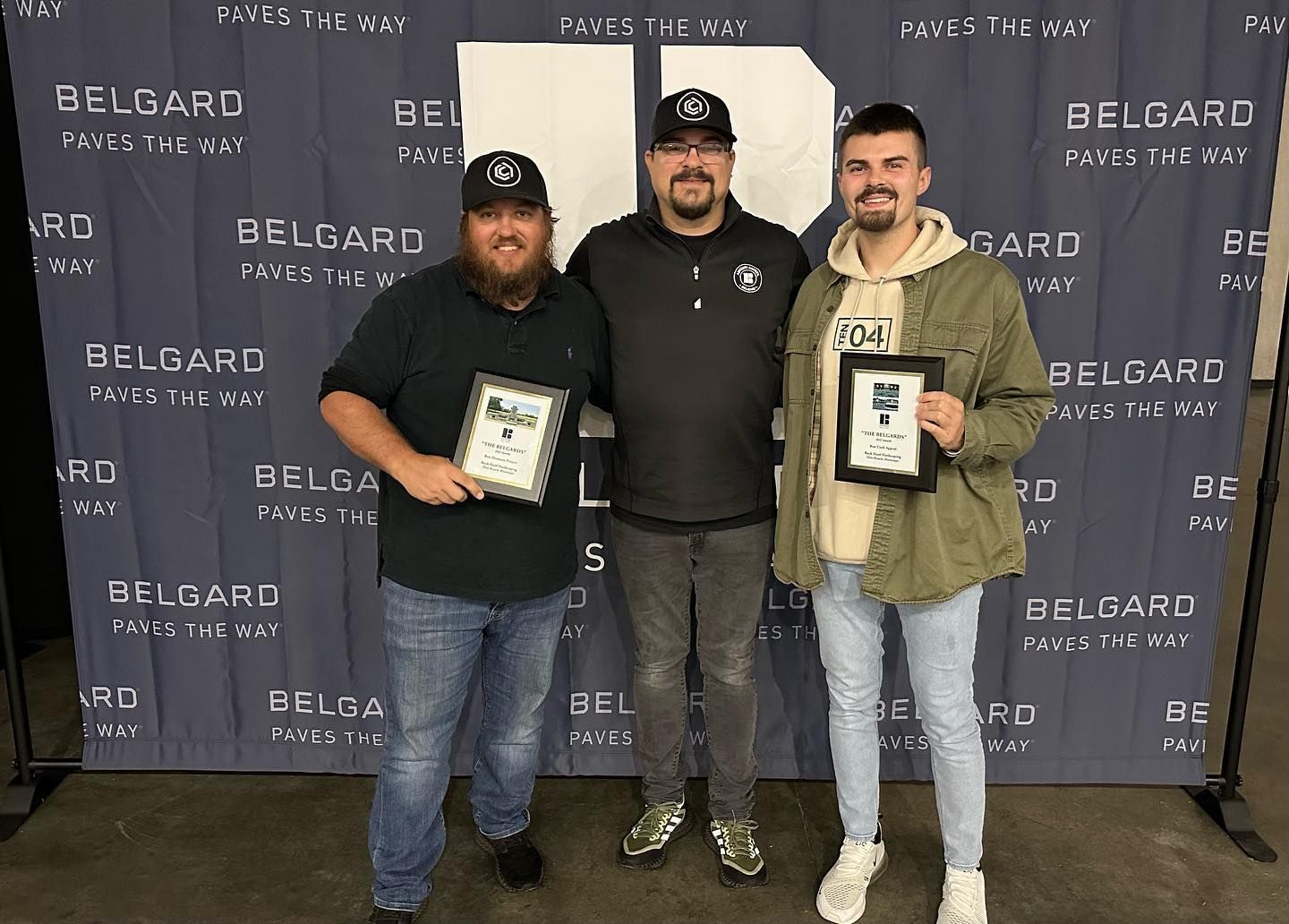 Three men holding plaques, posing in front of a blue Belgard banner. Two wear baseball caps.